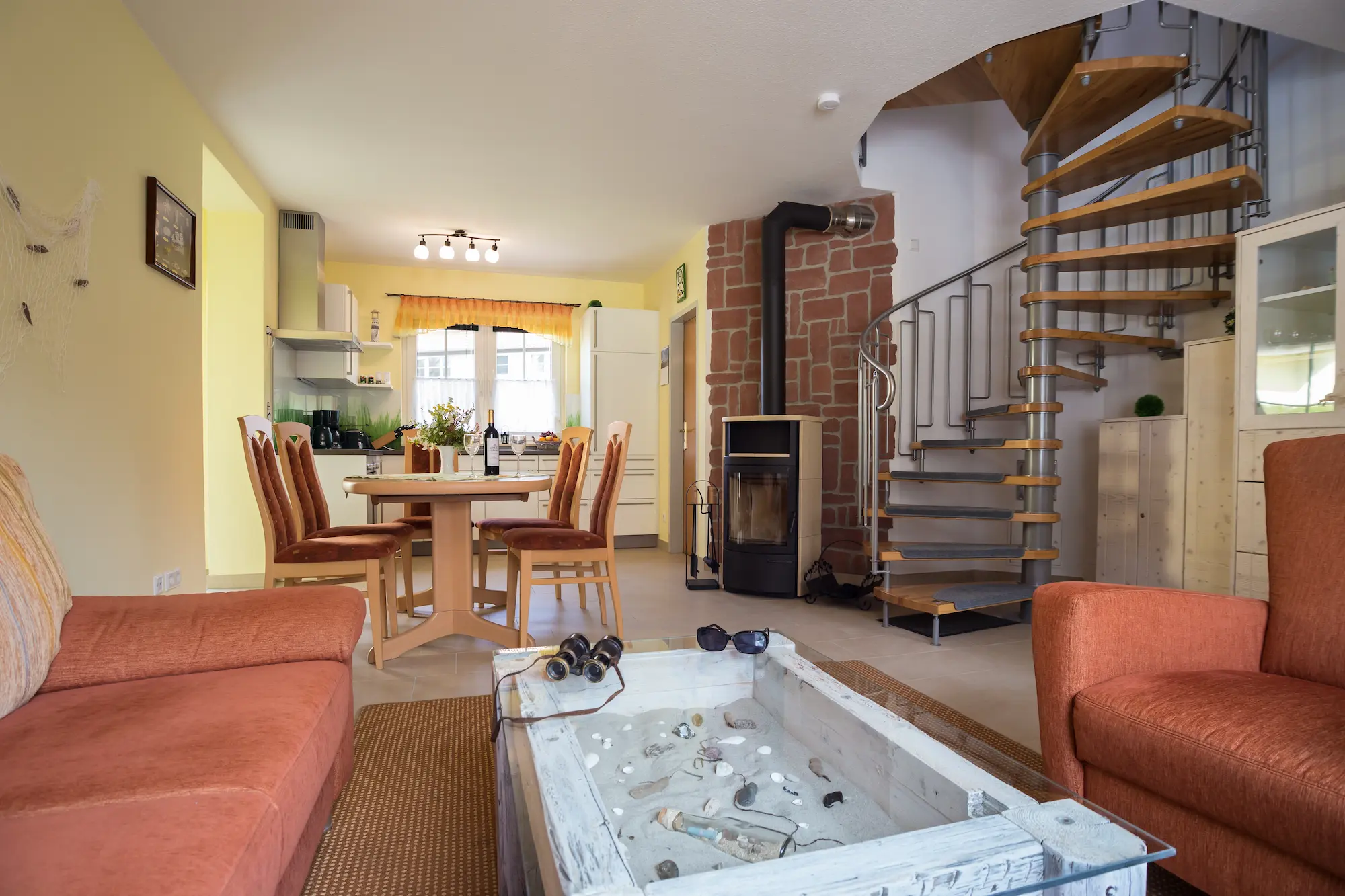 Offener Wohn- und Essbereich im Ferienhaus Am Deich 25 mit Sofa, Esstisch, Wendeltreppe und Blick zur Küche.