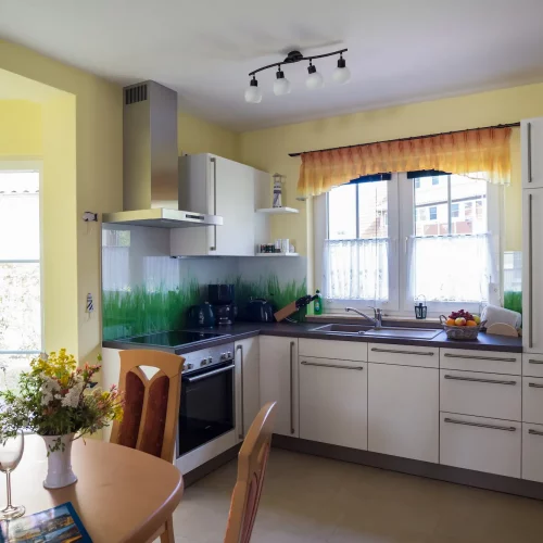 Helle Küche im Ferienhaus Am Deich 25 mit großem Fenster, Arbeitsfläche und Esstisch im Vordergrund.